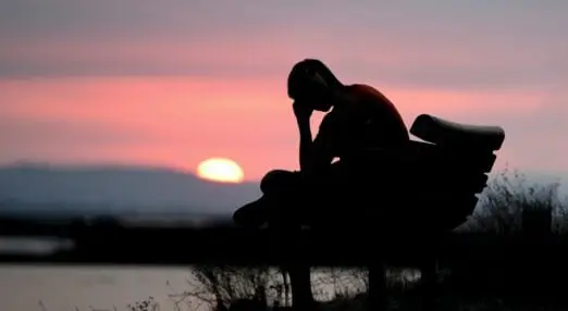 man sitting on a bench, head in hands, while the sun sets