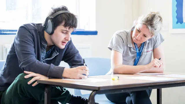 boy with headphones on writing at a desk supervised by a teacher