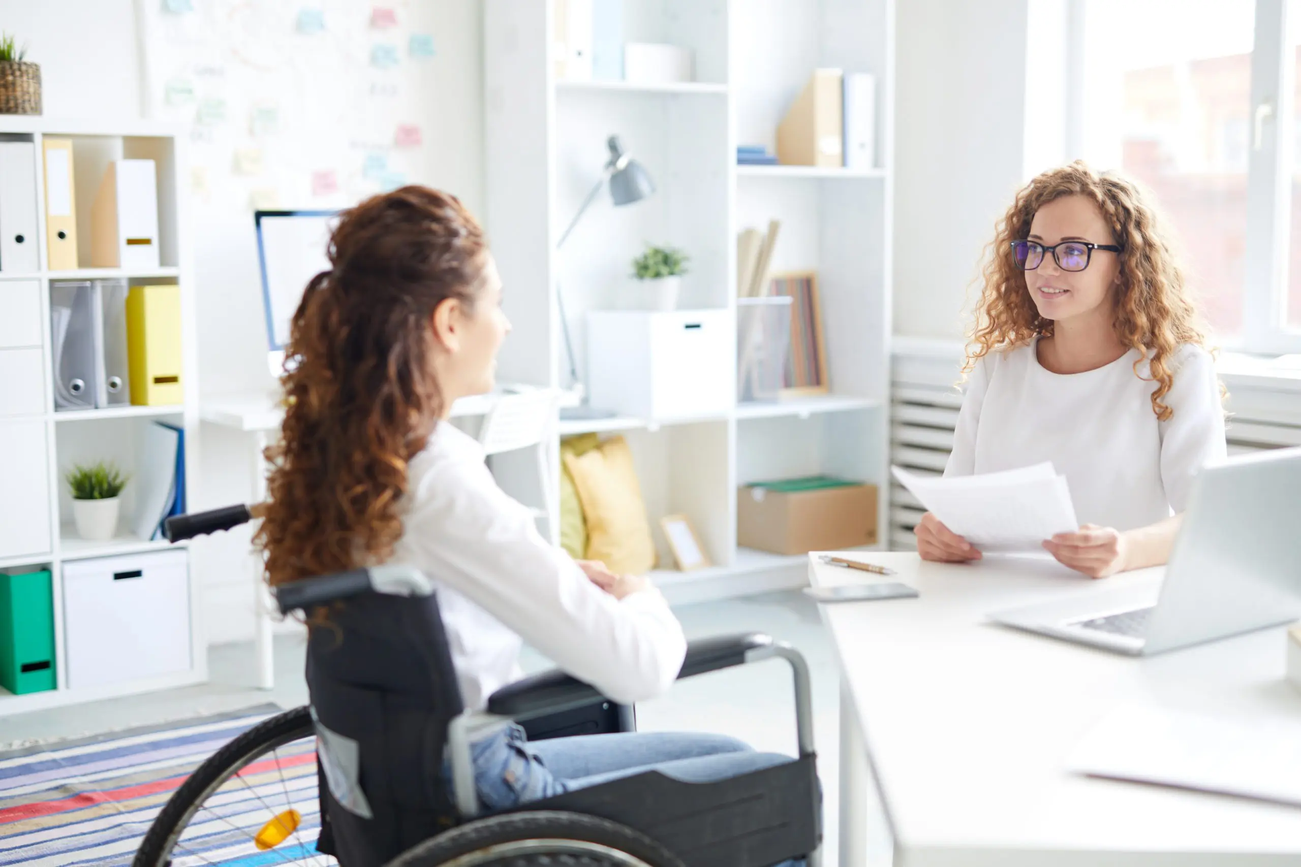 Two women talking to each other, one sits in a wheelchair, the other sits holding a piece of paper