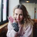 White woman in a pink jumper, holding a phone with a pink case looking into the camera