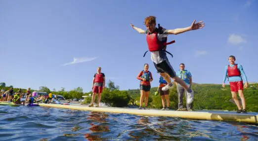 Young person jumping off a floating dock into the water on a sunny day, with others in life jackets nearby and lush green hills in the background.