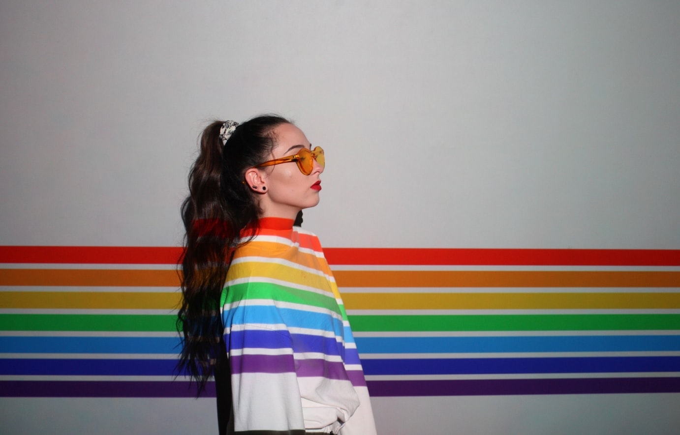 Profile of a girl with long hair and glasses wearing a white jacket with rainbow stripes on, blending into a rainbow stripe background.