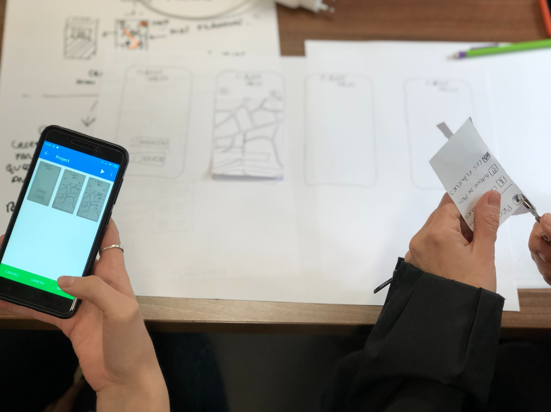 one person cutting out prototype drawings and another person holding a phone with photos of the prototype