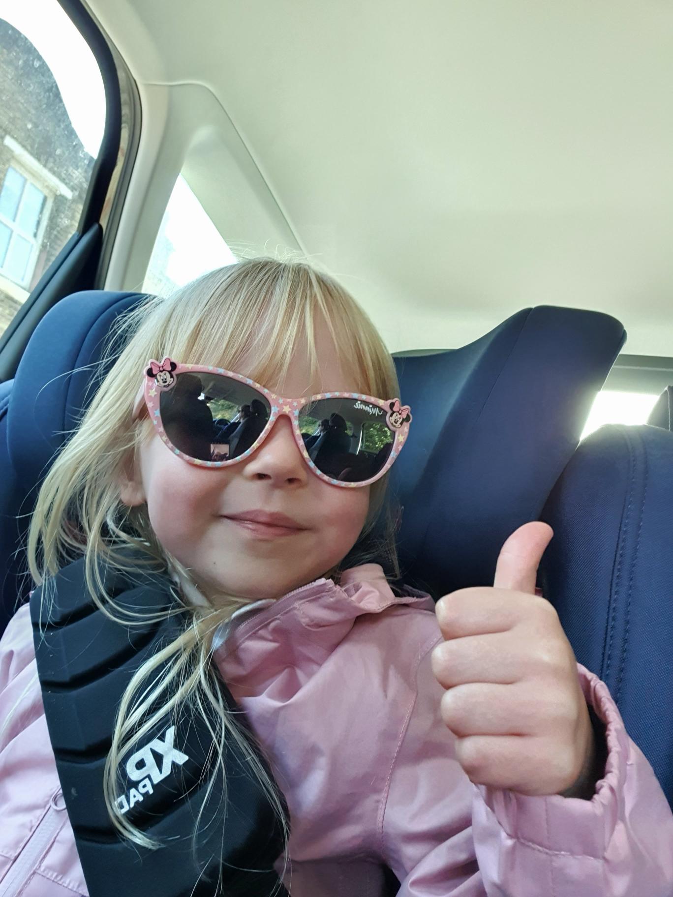 Blonde chid wearing sunglasses, sat in the seat of a car with thumbs up, smiling at the camera