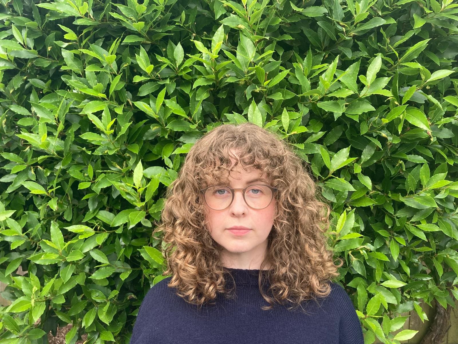 A white woman in glasses with curly blond hair wearing a dark blue top standing in front of leaves.